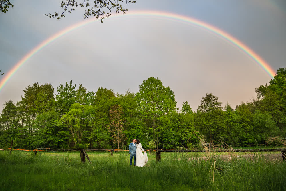 Regenbogen über der Wiese am Hof, das Brautpaar in der Ferne
